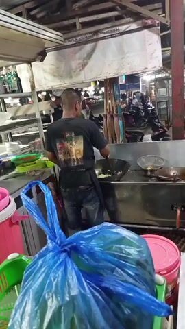 A man is cooking in an outdoor kitchen area at night. He is wearing a black t-shirt with a graphic on the back and dark-colored jeans. He stands in front of a large metal wok or frying pan, stirring its contents with a spatula. Behind him, motorcycles are parked along the street. The kitchen area is cluttered with various containers, dishes, and a metal shelving unit. In the foreground, a large blue plastic bag is tied off, and a green basket and pink bucket sit on the counter. A sign with text in Indonesian is visible above the cooking area.