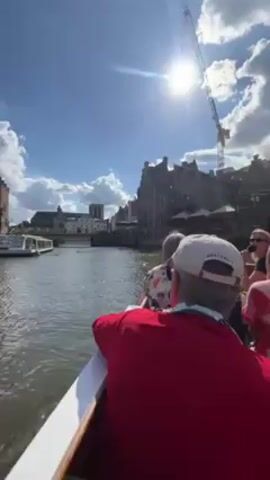 The video shows a boat tour on a river in a city. The camera is positioned on the boat, facing forward. In the foreground, the back of a man in a red shirt and a white cap can be seen. Behind him, other passengers are partially visible. The boat is moving through a canal or river with buildings lining both sides. The buildings are old and appear to be made of brick with distinctive architectural details. Several boats are docked along the riverbank. In the distance, a bridge and a tower can be seen. The sky is blue with scattered white clouds, and the sun is shining brightly, creating a glare. A construction crane is visible in the background.