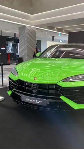 A vibrant lime green Lamborghini Urus SE is the central focus of this video, prominently displayed indoors, likely within a modern shopping mall or exhibition space in Daegu, South Korea. The luxury SUV is presented on a dark carpeted platform, with its distinctive front fascia and hood facing the camera. The car features contrasting black trim elements on its lower bumper, grille, and air intakes, complementing its bright color. A white display sign in front of the vehicle clearly identifies the model as "URUS SE" in black lettering.

In the background, a woman dressed in dark attire can be seen walking or standing behind the car's left side. The indoor setting is brightly illuminated with modern overhead lighting and features contemporary architectural details, including fluted pillars. Glimpses of other retail establishments are visible, with a "TUDOR" watch store sign clearly discernible in the left background, indicating a high-end commercial environment. The scene suggests a public showcase or launch event for the Lamborghini Urus SE.