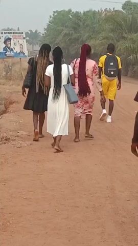 The video shows a group of young adults walking down a red dirt road. There are three women and two men. The women are wearing dresses and carrying handbags. The men are wearing casual clothes. The road is lined with trees and bushes. There is a sign on the left side of the road with a picture of a man on it. The sky is overcast.