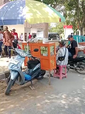 The video shows an outdoor scene with a food cart attached to a scooter. The cart is orange and has signs that say "RUJAK BUAH" and "IRIS". There are several people in the background, some standing and some sitting. A man is sitting on a pink stool next to the food cart, and another man is sitting on a scooter behind the cart. The ground appears to be dirt and gravel. There is a yellow and green umbrella providing shade over the food cart.
