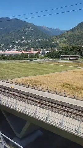 A train travels along a track next to a large green field and a dry golden field. In the distance, mountains rise behind a town with buildings. The sky is clear and blue. A tall utility pole is visible next to the train tracks. The camera is looking out from inside the train.