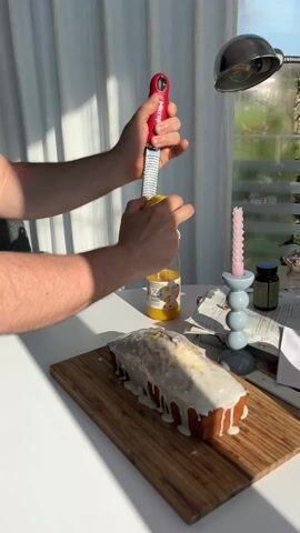 A person zests a lemon with a Microplane grater over a loaf cake that sits on a wooden board. The cake has a white, dripping glaze. In the background, there is a window with a white curtain and a silver lamp. Other items on the table include a bottle of orange juice, a pink candle in a light blue candlestick, and a bottle of pills. The person's hands are visible holding the lemon and grater, with a wedding band visible on one finger.