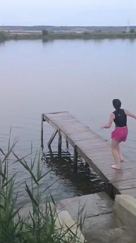A young woman with dark hair, wearing a black tank top and pink shorts, runs towards the end of a wooden pier that extends over a body of calm water. She leaps off the pier, performing a dive into the water, causing a splash. A young girl in a light-colored shirt and dark shorts stands on the steps leading down to the water, observing the dive. The environment is natural, with tall grasses in the foreground and a treeline in the background under an overcast sky.