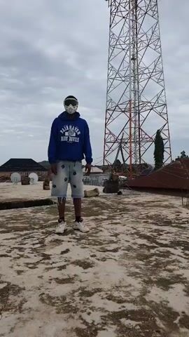 A person wearing a blue hoodie, denim shorts, and a mask is doing push-ups on a rooftop. A tall metal tower stands in the background. The sky is cloudy.