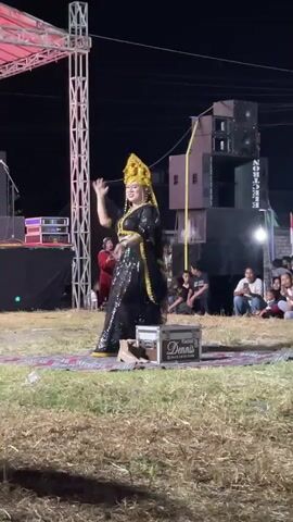 A woman in traditional attire with a golden headdress and black and gold sequined dress is dancing on a rug in front of a stage. She is wearing a black veil and her dress has a long skirt that flares out as she moves. There are speakers and lighting equipment behind her. An audience is seated in the background. The event appears to be outdoors at night.