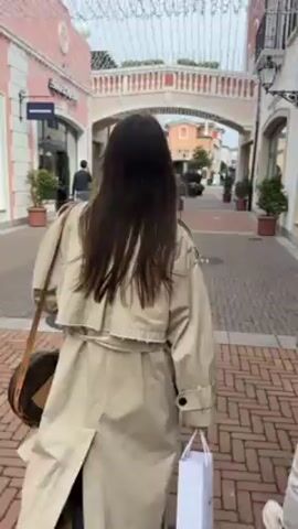 Two women stroll down a brick-paved street lined with shops. The woman in the foreground wears a beige trench coat and a brown purse, while the other person is partially visible to the right. The shops on either side of the street have a European architectural style with archways and potted plants. Decorative lights are strung overhead. Several other people can be seen walking in the distance.