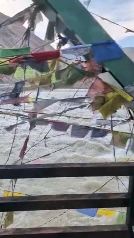 The video shows a view of a fast-flowing river from what appears to be a bridge or elevated platform. Prayer flags are strung across the foreground, fluttering in the wind. The river is wide and turbulent, with white water indicating a strong current. In the background, steep, mountainous terrain is visible under a cloudy sky. The overall scene suggests a remote, possibly high-altitude location.