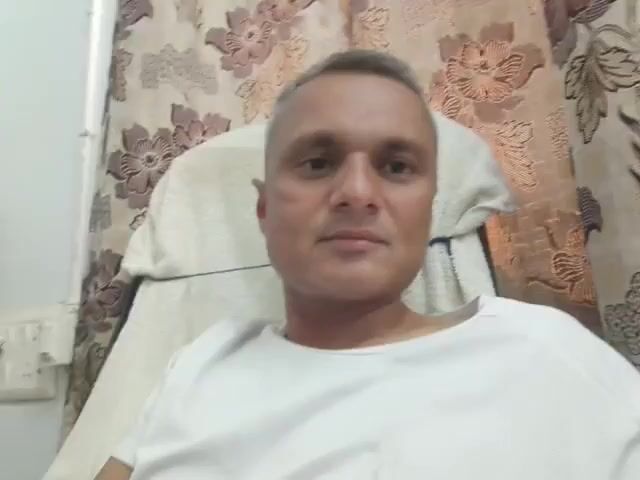The video shows a close-up, head-and-shoulders shot of a man with short, graying hair. He is wearing a white t-shirt and is seated in a chair with a light-colored blanket draped over it. The background features floral patterned curtains. The man is looking directly at the camera and appears to be speaking, although no audio is present. His facial expression is neutral at the beginning of the clip and transitions to a slight smile towards the end. There is a visible scar on his left cheek. A light-colored electrical outlet with multiple sockets is visible on the left side of the frame.