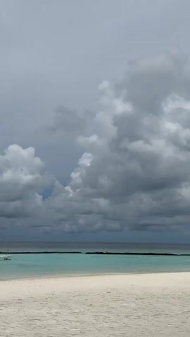 This video shows a tropical beach scene under a cloudy sky. The foreground features a white sand beach. Beyond the beach, the ocean is a vibrant turquoise color, separated by a dark line in the water, possibly a submerged barrier or sandbar. In the distance, a small boat with a white canopy is visible on the water. The sky is filled with dramatic gray clouds, with a hint of blue sky visible in the upper center. On the left side of the frame, a palm tree leans over the beach, and a small airplane can be seen flying in the distance. The overall mood is serene, with a hint of impending weather due to the clouds.