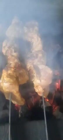 A close-up shot shows chicken drumsticks skewered and roasting over glowing coals. Smoke rises from the meat as it cooks, with visible charring and glistening skin. The chicken pieces are arranged closely together, some overlapping. The camera pans slightly to reveal more skewers of chicken, creating a dense arrangement of food being grilled. The lighting is dim, with the red glow of the coals providing an undercurrent of light.