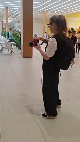 A woman with blonde hair pulled into a ponytail, wearing a white t-shirt, black vest, and black pants, stands in a brightly lit indoor space with a tiled floor and a modern ceiling with strip lighting. She is holding a red camera with a lens attachment and appears to be filming. She has a black backpack on her right shoulder with some decorations attached. In the background, there are tables and chairs, plants, and other people, including a woman in a long white dress. The woman with the camera turns and walks away from the viewer.