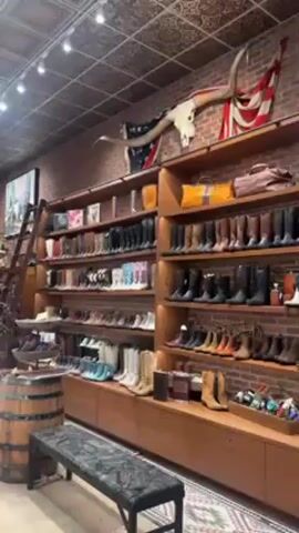 The video shows the interior of a high-end clothing and accessories store. The store has a rustic decor, with exposed brick walls, a wooden ceiling, and vintage-style lighting. There are shelves filled with cowboy boots in various colors and styles, as well as clothing racks with jackets and other apparel. There are also display cases filled with jewelry. One person is visible in the video, walking around the store and browsing the merchandise. The overall impression is that the store specializes in Western-themed luxury goods.