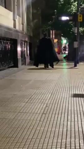A person dressed as Batman walks away from the camera down a city sidewalk at night. The sidewalk has a striped pattern.  Other people are visible in the distance. Buildings line the street. A red post box is also visible. The Batman figure wears a black cape.
