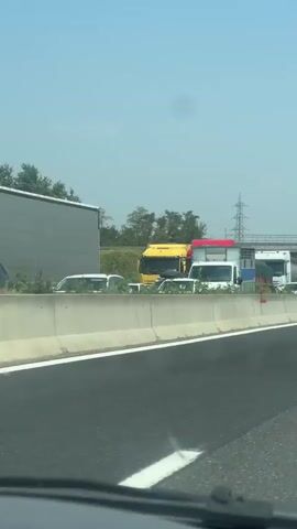 A traffic jam is visible on a highway under a blue bridge. Several trucks and cars are stopped on the road. The weather appears to be clear and sunny. The scene is viewed from inside a vehicle on the highway.