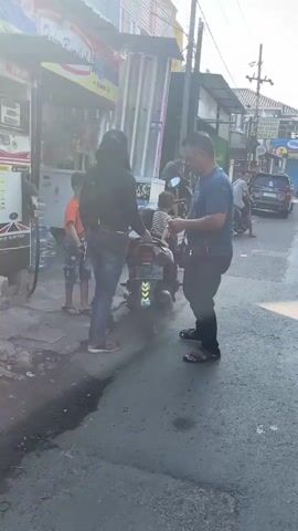 The video shows a street scene with several people and a parked motorcycle. In the foreground, two men and a young boy are interacting near a parked motorcycle. One man, wearing a blue t-shirt and dark pants, is holding a small object, possibly a phone or wallet, in his hands and looking down at it. He appears to be engaged in a transaction or conversation with the person next to him. The second person, whose face is obscured by a dark hooded jacket, is standing by the motorcycle and reaching towards the man in the blue shirt. A young boy, wearing an orange t-shirt, stands to the left of the motorcycle, facing away from the camera.

Behind the motorcycle, another person, possibly a child, is partially visible sitting on the motorcycle. The environment appears to be an urban street, with buildings visible on the left and right sides of the frame. There is a structure on the left that looks like a small shop or stall, possibly a convenience store, with items displayed. The ground is paved and appears wet in places, suggesting recent rain or cleaning. In the background, other vehicles, including a car and a motorcycle, are visible on the street. The lighting suggests it is daytime, with sunlight casting shadows. The overall atmosphere seems to be a casual street encounter.