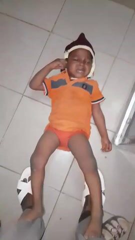 A young child is lying on a tile floor, wearing an orange shirt, orange underwear, and a burgundy cap with a white band. The child is crying, thrashing their arms and legs, and attempting to crawl.  A person standing nearby is wearing gray pants and white sandals. The person reaches down and briefly touches the child.