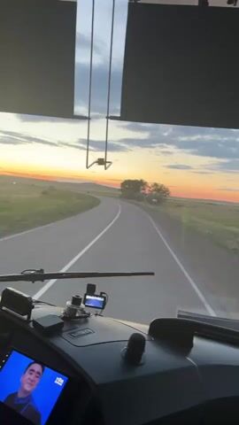 The video begins with a view from the inside of a vehicle, looking out the windshield. The dashboard is visible in the foreground, featuring a screen displaying what appears to be a music video or slideshow. The landscape outside consists of a paved road curving through a grassy, rural area. In the distance, a sunset paints the sky with warm colors of orange, yellow, and pink, with clouds scattered across the horizon. Trees and hills are silhouetted against the colorful sky. The vehicle is moving forward, as indicated by the blurring of the road and the perspective. The camera angle suggests the footage is shot from the driver's or passenger's seat. The overall scene conveys a sense of travel through a scenic, natural environment during sunset.