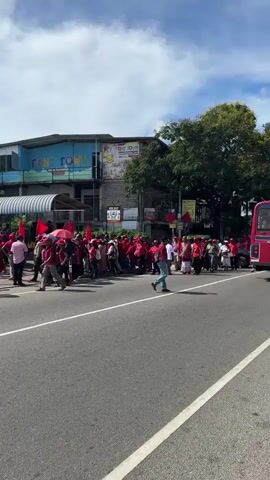The video shows a group of people, most wearing red shirts, gathering on the side of a road. Many of them are holding red flags. The location appears to be an urban area with a building that has "TINY TOWN" written on it. A person is walking across the road in front of the group. The sky is partly cloudy.