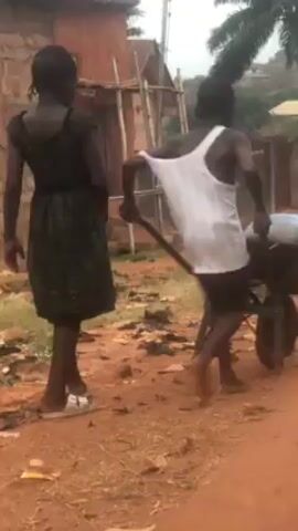 The video shows two individuals on a dirt road. One is a young person in a black dress and white shoes, walking next to another young person who is pushing a wheelbarrow. The person pushing the wheelbarrow is wearing a white tank top and dark shorts and appears to be barefoot. The wheelbarrow contains a blue container. The road is made of red dirt, and there's scattered debris on the ground. In the background, there's a partially constructed structure with scaffolding, some vegetation, and distant buildings. The video seems to be taken in a somewhat rural or developing area.