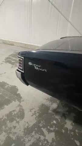 The video shows the side and rear of a dark-colored car, possibly blue or black. The car has "Super Sport" badging on the rear quarter panel and is parked indoors on a concrete floor. The car has silver wheels with white-lettered tires that say "Cooper Cobra Radial G/T". The interior of the building appears to be a large, enclosed space with white, tent-like walls and visible structural supports. Tire marks are visible on the concrete floor.