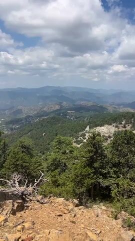 The video shows a panoramic view of a mountainous landscape with a village nestled in the valley below. The foreground is dominated by rocky terrain and trees, while the background consists of rolling hills and mountains stretching into the distance. The sky is partly cloudy. The village appears to be a cluster of buildings with red roofs surrounded by lush green vegetation. A winding road is visible on the right side of the frame, leading through the trees. The overall impression is one of natural beauty and rural tranquility.