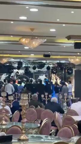 The video shows a gathering in a large hall, likely a reception or wedding. Many people are present, some seated at round tables adorned with centerpieces and elegant chairs, while others are congregated near a raised platform that appears to be a stage. This area is decorated with white fabric or flowers and is equipped with lighting fixtures and sound equipment. The hall is well-lit with recessed lights and ornate chandeliers. The overall atmosphere suggests a formal or celebratory event.