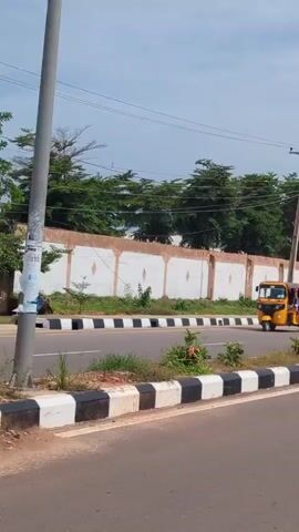 This media file, captured in Unguwar Rimi, Nigeria, presents a daytime scene on a paved road. The weather appears partly cloudy, with a bright sky suggesting it's likely late morning or afternoon.

The primary subjects are vehicles, including a yellow auto-rickshaw with passengers and a motorcycle. Several buildings are visible in the background, ranging from modern multi-story structures to what appear to be residential homes with tiled roofs. A long, light-colored wall with a decorative border runs alongside the road, separating it from the properties behind.

The scene depicts everyday traffic and activity on a road. The presence of streetlights, one with solar panels, suggests infrastructure development. The road itself is clearly marked with black and white striped borders, indicating a well-maintained public area.

The overall impression is one of a typical day in a developing urban or semi-urban area. There are no strong emotional cues evident, and the focus is on the environmental and vehicular elements. No discernible text is visible within the frame.