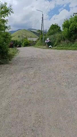 The video shows a dirt road with green trees and rolling hills in the background. There is a pole with wires and a street light on the right side of the road. A garbage can is also visible next to the pole. In the distance, a red vehicle is parked on the left side of the road. The sky is partly cloudy.