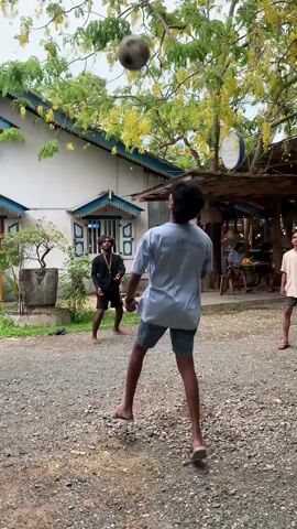 Three young men are playing sepak takraw in a gravel clearing between a building and an open-air structure with a sign saying "The Hideout". The building is two-story, white, with teal trim on the windows and roof. The sky is overcast. The men are wearing t-shirts and shorts and are barefoot or wearing flip-flops. One man serves a woven ball, and another kicks it. Another man is watching them. As the ball is kicked toward the right of the frame, two of the men follow it in that direction.