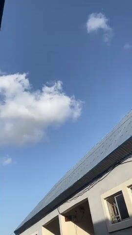 This media file shows an upward-facing shot of a building under a bright blue sky with scattered white clouds. The building appears to be a single-story structure with a light gray facade. It has a sloping metal roof, likely blue or dark gray, with visible seams and fasteners. A window with a white frame is visible on the right side of the building, showing some items inside, possibly bottles or containers. Below the roofline, there are several recessed openings that appear to be entrances or covered areas. Electrical conduits and a junction box are attached to the exterior wall.

The sky is a clear, vibrant blue, indicating a sunny day. The clouds are puffy and white, suggesting fair weather. The lighting suggests it is daytime, likely late morning or early afternoon due to the brightness and the angle of the sun, which is not directly visible. The image does not show any people or specific activities taking place. The presence of electrical infrastructure and the style of the building suggest a residential or commercial area, possibly in a tropical or subtropical climate.

The location context provided is Pantinapu, Nigeria. This context, combined with the visual cues, points to a scene within Nigeria. There is no visible text in the media file. The overall mood conveyed is one of a bright, ordinary day.