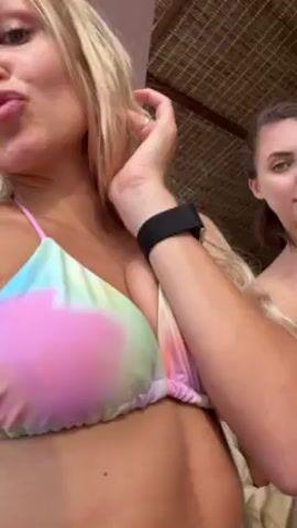 Two young women are in what appears to be a resort. One woman, closest to the camera, has long blonde hair and is wearing a tie-dye bikini top and denim shorts. She's holding the camera and smiling, sticking out her tongue, making a hang loose gesture, and laughing. The other woman has brown hair pulled back and is wearing a white strapless top and shorts. She is holding a phone and posing, sticking out her tongue, making a rock-on gesture, and laughing. There are plants and chairs visible in the background, and music is playing.
