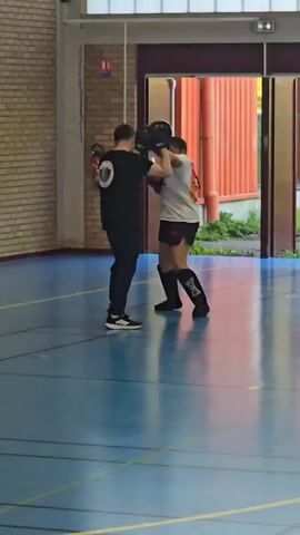 The video shows two people practicing martial arts indoors. The room has a blue floor with navy lines, and the walls are light yellow. A man in a black shirt, black pants, and Adidas sneakers stands facing a woman. He is holding two black padded targets. The woman wears a white t-shirt, black shorts, black shin guards, and red boxing gloves. She is practicing a kick towards the targets held by the man. There is a doorway behind them, leading to a building with red corrugated metal siding. There is some greenery visible outside the doorway.