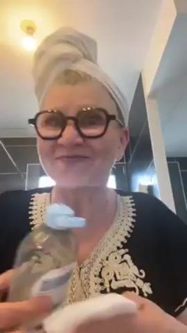 A woman is in her bathroom, seen from the chest up, wearing a black patterned robe and round-framed eyeglasses. She has a towel wrapped around her head. With an unimpressed expression, she pours a clear liquid from a blue-capped bottle onto a white cotton pad held in her other hand. A lightbulb is visible on the ceiling above her. The bathroom has black and grey tiles.