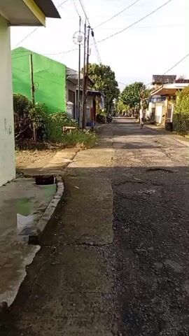 A person in a light-colored shirt and dark pants walks down a narrow asphalt road lined with houses and greenery. The road appears to be in disrepair with cracks and patches. Sunlight casts shadows, indicating it is likely daytime. The houses are simple structures, some with laundry hanging outside. A motorbike is parked on the side of the road.