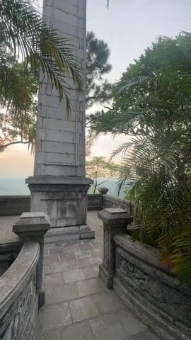 This media file shows a stone balcony with ornate railings and a tall, fluted stone column. Palm fronds and other lush greenery surround the balcony, suggesting a tropical or subtropical climate. Beyond the railing, a scenic view unfolds of a rolling landscape with trees and a hazy sky, possibly indicating dawn or dusk due to the soft, diffused light. The paving stones on the balcony are also made of stone, laid in a patterned arrangement. The overall impression is one of an ancient or historical site, perhaps a temple or monument, offering a vantage point for observing the surrounding natural beauty. No people are visible. The time of day appears to be either sunrise or sunset, judging by the warm, soft light and the sky's hue. The location context provided places this scene in Phong Châu, Vietnam, which aligns with the presence of lush vegetation and potentially historical architecture. The mood is serene and contemplative. There is no visible text in the media.