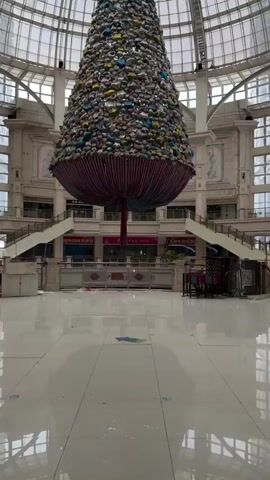 This video shows a large, conical structure resembling a Christmas tree, suspended from the ceiling of a grand, indoor atrium. The tree is densely covered in what appear to be numerous bras in various colors, including pink, yellow, blue, and grey, with a golden star at its apex. The lower section of the tree is adorned with red and white striped fabric.

The atrium features a domed glass ceiling, allowing natural light to filter in. The space is multi-leveled, with grand staircases leading to upper floors. The architecture suggests a large public building, possibly a shopping mall or a commercial center, given the presence of shop fronts visible on the lower levels. There is some visible text on signs behind the tree, but it is not clearly legible.

The scene is devoid of people, creating a sense of stillness and perhaps emptiness. The floor is a large, polished white tiled surface, reflecting the light and the suspended tree. The overall atmosphere is somewhat surreal due to the unusual material used for the tree's decoration. The time of day is indeterminate due to the indoor setting, but the lighting suggests it is daytime. The weather outside is not visible. The location is identified as Guta, China.