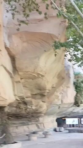 The video begins with a close-up view of a sheer rock face, showcasing its layered texture and weathered appearance. As the camera pans to the right, a large tree with dense foliage comes into view, its branches extending over a paved area. Several picnic tables with stone bases are scattered around the base of the rock face. Further to the right, a group of people are visible in the distance, appearing to be walking or standing near some informational displays. The environment suggests an outdoor recreational area, possibly a park or a natural attraction. The lighting is somewhat diffused, indicating it might be late afternoon or early morning. The overall scene is serene and natural.