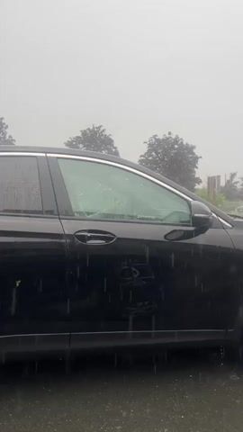 A black SUV is parked on a wet asphalt surface, likely a road or parking lot, during a rain shower. Water droplets are visible hitting the car and the ground. The car's windows are reflecting the overcast sky and trees. In the background, more trees with dark foliage and other vehicles are visible through the rain. The sky is a uniform gray, indicating heavy cloud cover. There are no people clearly visible inside or outside the vehicle. The camera is positioned at a medium distance, focusing on the side of the SUV.