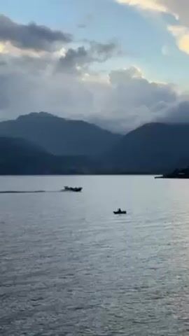 The video shows a body of water with mountains in the background. The sky is partly cloudy, and the light suggests it might be dawn or dusk. There are two boats visible on the water. One appears to be a larger boat moving at a moderate speed, leaving a trail behind it. The other is a small kayak or similar vessel with a single person on board, moving slower than the larger boat. The overall scene conveys a sense of tranquility.