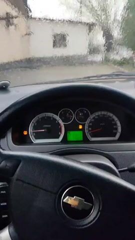 The video, shot from inside a car on a rainy day, shows the dashboard and windshield wipers in operation. The dashboard features a speedometer, a fuel gauge, and a digital display showing "P 1686." Outside the windshield, a small white building with a brown roof and a single window is visible. A leafless tree stands next to the building, along with two evergreen trees. Rain is pelting the windshield, and the wipers are clearing the water. The overall impression is one of a dreary, rainy day in a rural setting.