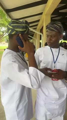 The video opens on two young men standing next to each other. The man on the left, facing away from the camera, is wearing a white traditional outfit and a striped cap. He holds a phone to his ear. The man on the right, facing the camera, is also wearing a white traditional outfit and a striped cap. He is holding a phone in his hand and appears to be speaking to the man on the left. Behind them, a crowd of people are seated on wooden benches under a covered outdoor structure with a slatted roof. The man on the right turns and walks away from the camera, moving further into the gathering. The camera follows him as he passes by the seated attendees. The video ends with a view of the man's back as he continues into the crowd.