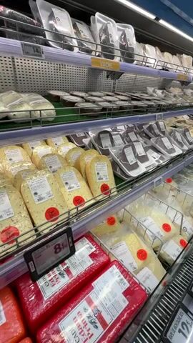 The video shows a wide aisle in a supermarket, featuring a display of various cheeses. The cheeses are packaged and arranged on shelves, with some sections organized by type, such as "Vache" (cow's milk cheese). The lighting is bright, typical of a retail environment. The camera pans along the shelves, showcasing the abundance and variety of cheese products. There are no people visible in the immediate vicinity of the cheese display, and no specific actions or interactions are depicted. The environment appears clean and well-organized.