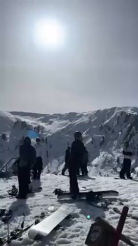 The video shows a sunny, snow-covered mountaintop. Several people are standing or sitting on the snow.  Some appear to be skiers or snowboarders. Ski equipment is lying on the ground. There is a blue flag with writing on it. In the background are snow-covered mountains and some trees. The sun is high in the sky.