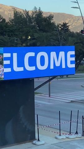 The camera captures a "WELCOME TO BUDVA!" digital sign in the forefront, displaying white lettering on a blue background. The sign is mounted on a dark metal structure, with crosswalk and city streets visible behind it. Cars of various colors pass by the sign, including a white car with green decoration and a gray hatchback. A pedestrian wearing gray clothes and a cap crosses the street behind the sign, appearing to talk on a mobile phone. In the background, a line of trees and mountains can be seen under a clear sky. The scene suggests an outdoor urban environment, likely in Budva, Montenegro. The overall lighting suggests it is daytime. A red and white striped barrier is partially visible at the bottom of the frame, suggesting ongoing construction or maintenance nearby.