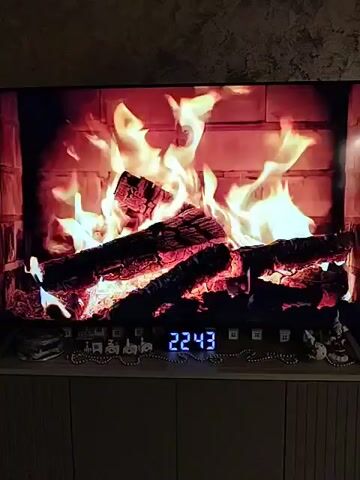 The video shows a living room decorated for the holidays. A large TV displays a fireplace scene with flickering flames and burning logs. The TV sits on a light-colored cabinet adorned with small decorative figurines and a digital clock displaying 22:43. To the left of the TV is a large black speaker with blue lights. Further to the left stands a Christmas tree decorated with white and silver ornaments, as well as small white lights. A decorative sock hangs on the wall next to the speaker. The overall environment suggests a cozy, festive atmosphere in a home.