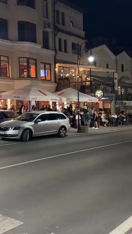 The video pans across a street scene at night. In the foreground, a silver station wagon is parked on the left side of the road, facing away from the camera. Across the street, a row of buildings with outdoor seating areas are visible. Several people are seated at tables under white umbrellas, some of them interacting and appearing to be dining or socializing. The buildings are illuminated, and a neon sign is visible on one of them. Streetlights line the road, and a few cars are seen driving in the distance. The overall impression is of a lively, urban environment at night, with people enjoying outdoor dining and nightlife.