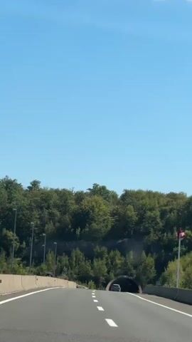A car is driving on a highway towards a tunnel. The sky is clear blue and the sun is shining. The highway is paved and has white lines marking the lanes. Trees and vegetation line the sides of the highway. The tunnel is dark and has a curved entrance. The car is the only vehicle visible in the shot.