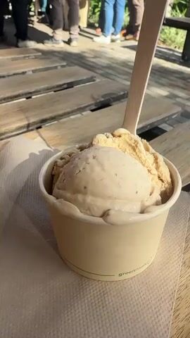 The video shows two scoops of ice cream in a paper cup with a wooden spoon inserted. The cup is placed on a napkin on a wooden picnic table. In the background, a line of people are standing and walking, some are wearing casual clothes. The setting appears to be outdoors, possibly a park or an outdoor cafe, with sunlight casting shadows on the table.