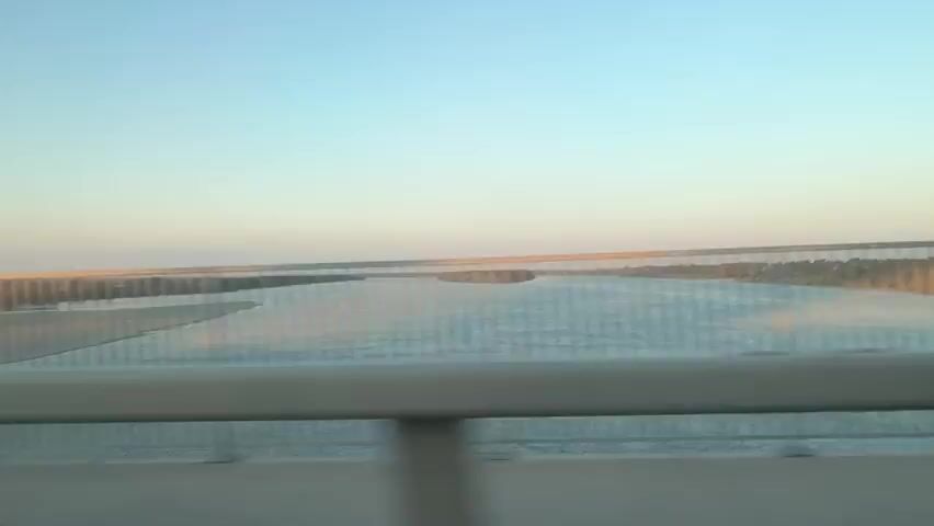 The video shows a view from a moving vehicle on a bridge, looking over a wide body of water with land on either side. The sky is clear and blue. The sun appears to be low in the sky, possibly indicating early morning or late afternoon. The water surface is calm, reflecting the light. There are no people visible. The vehicle passes by bridge railings and a light pole.